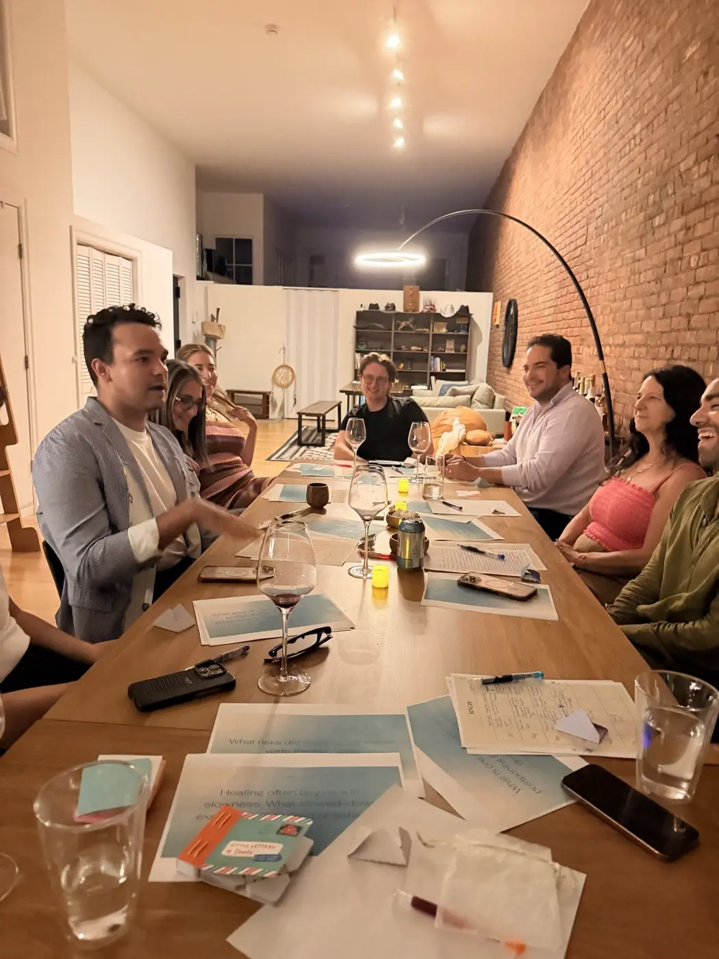 guests writing at a long wooden table under an arc lamp, with wine glasses and printed prompt cards, during a goal setting night in a SoHo living room, New York