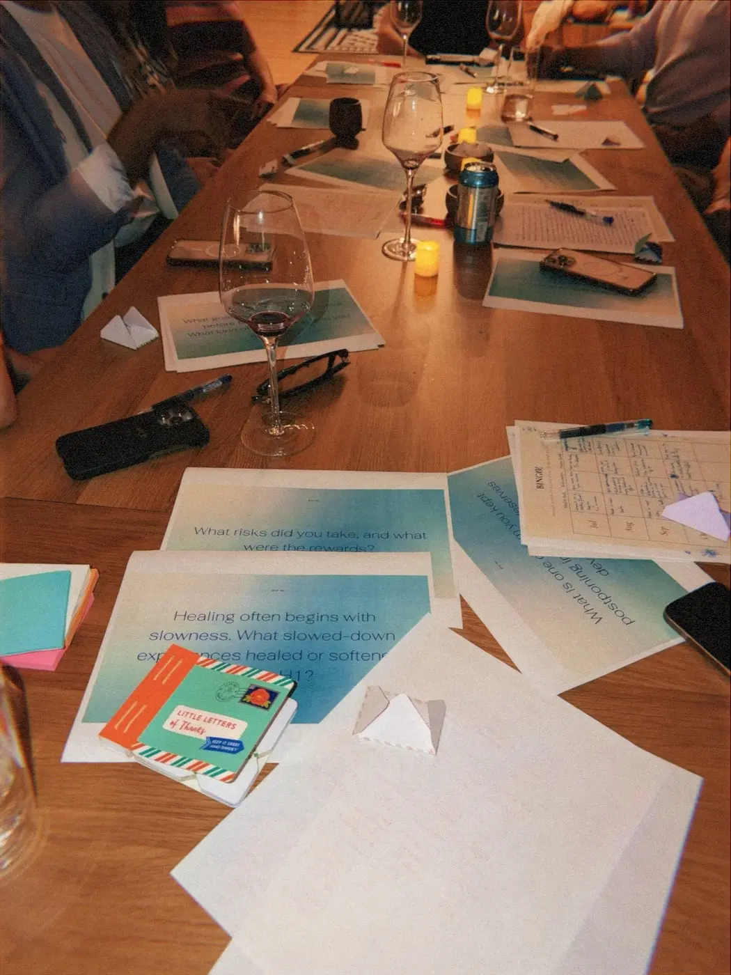 the long table mid evening, covered in blue prompt sheets, pens, wine glasses and a small envelope, during a goal setting night in a SoHo living room, New York