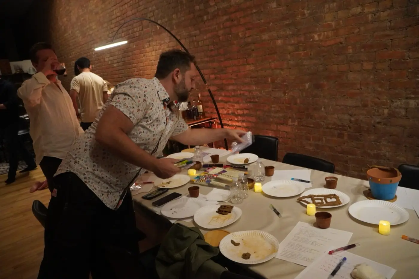 a guest handing out coloured markers across a long table of clay, paper plates and tealights, during an art therapy night in a SoHo living room, New York