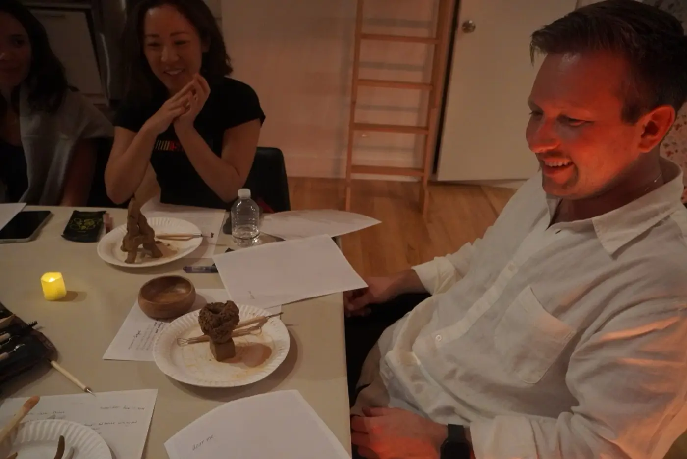 guests laughing beside small clay figures on paper plates and a prompt sheet with the words dear me printed at the top, during an art therapy night in a SoHo living room, New York