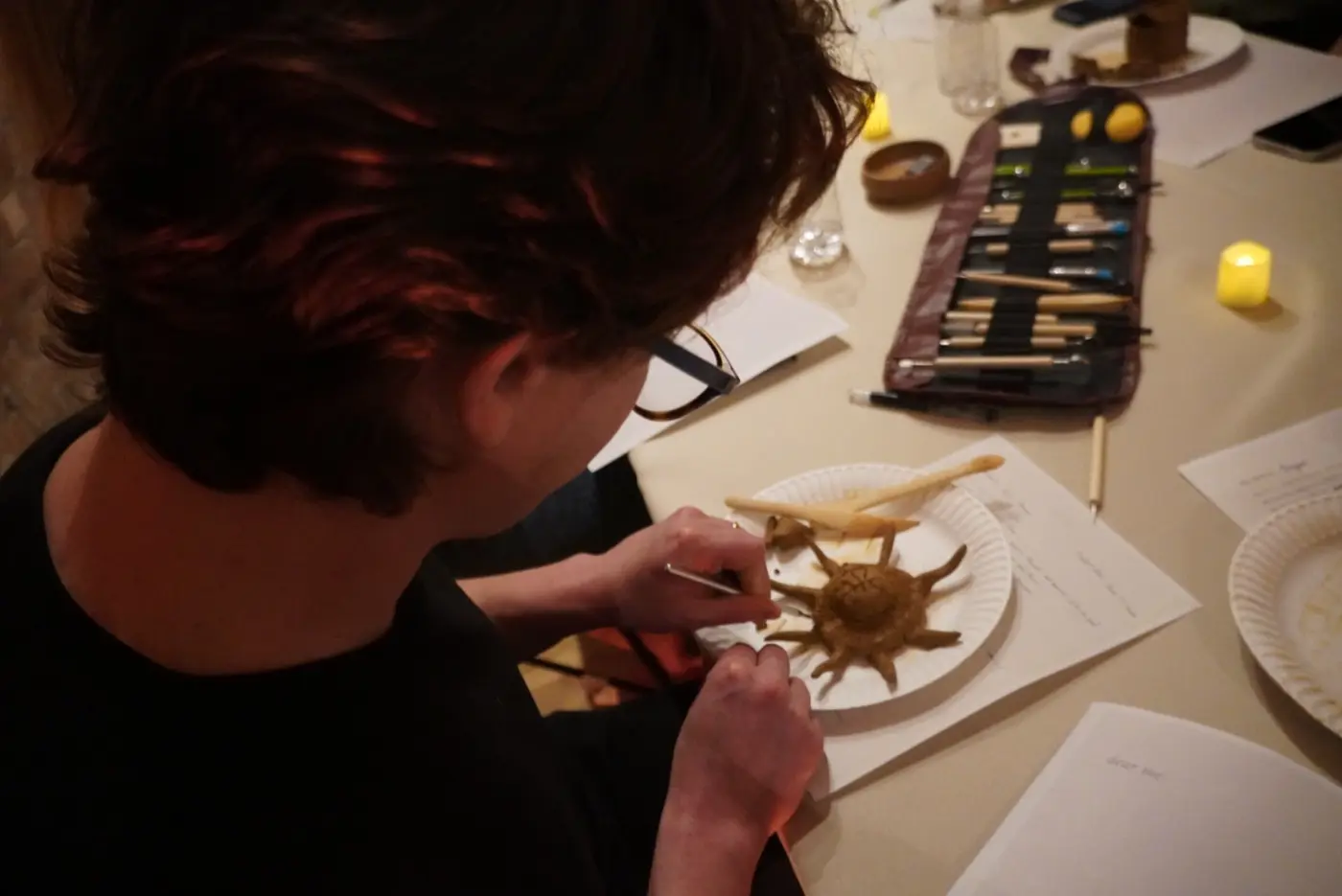 a guest shaping a clay sun on a paper plate beside a roll of sculpting tools and a dear me prompt sheet, during an art therapy night in a SoHo living room, New York