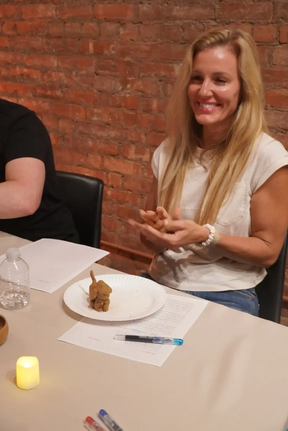 a guest laughing in front of an exposed brick wall with a small clay dog she has made on a paper plate, during an art therapy night in a SoHo living room, New York