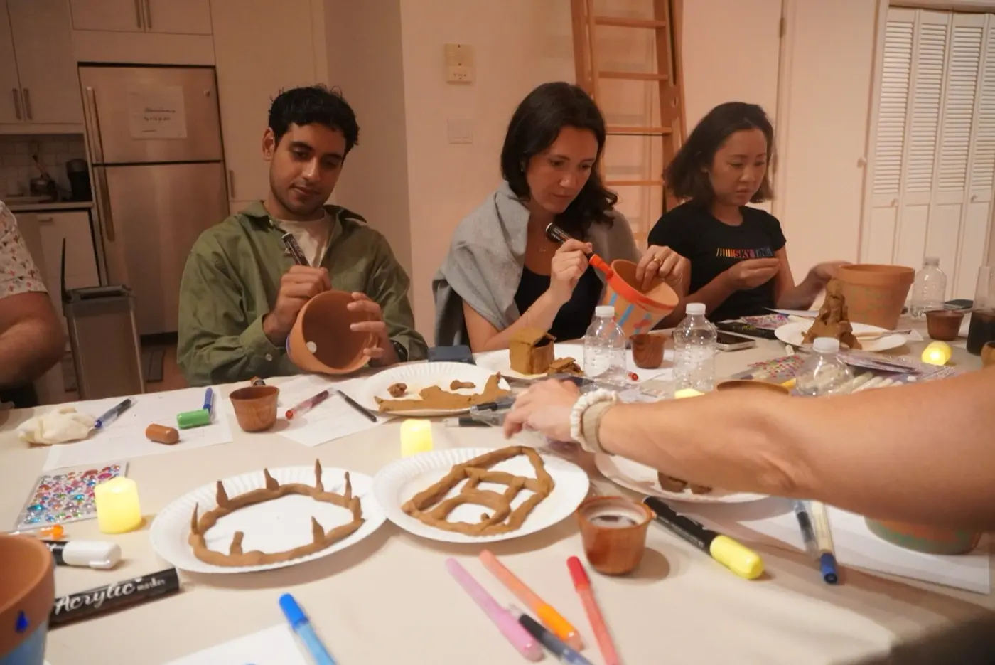 three guests painting terracotta pots with brushes and markers at the long table, during an art therapy night in a SoHo living room, New York