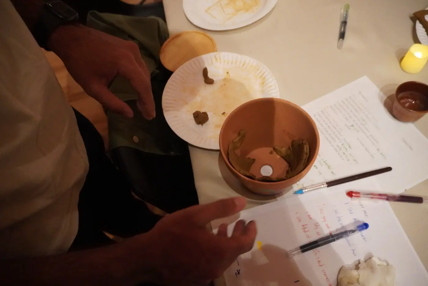 a guest pressing clay into a terracotta pot beside handwritten dear me sheets and coloured gel pens, during an art therapy night in a SoHo living room, New York