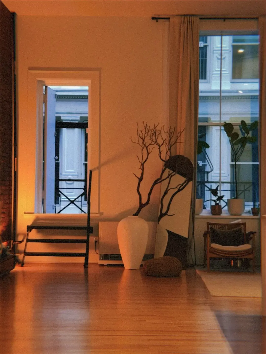 the living room at dusk lit warm, with bare branches in a tall white vase and a woven pouffe on the wooden floor, before a cacao ceremony in SoHo, New York