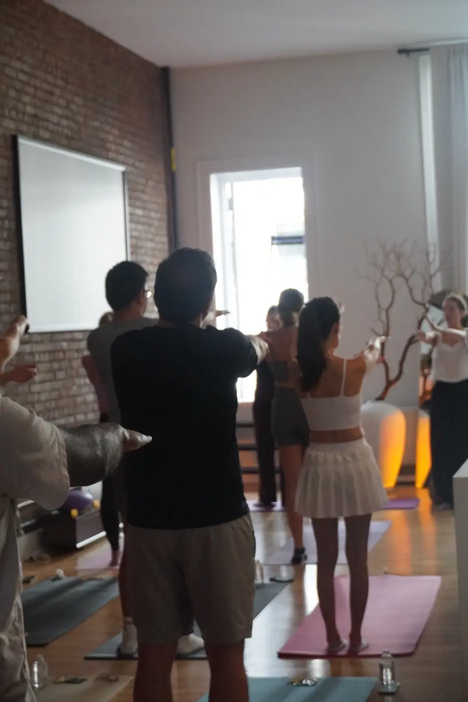 guests standing on mats with both arms stretched forward in front of an exposed brick wall, during a pilates class in a SoHo living room, New York