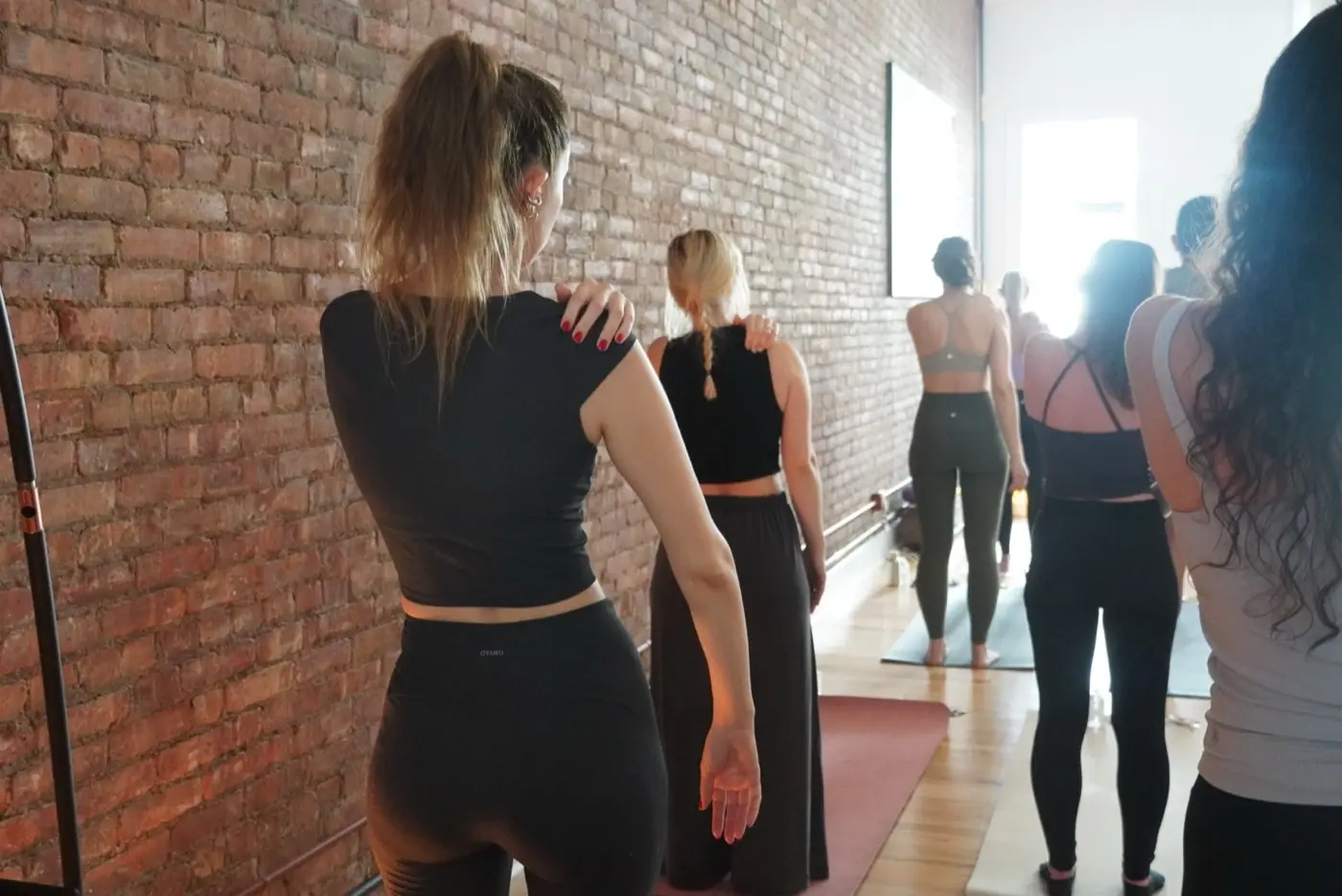 two women standing one behind the other with a hand on the shoulder in front of a whitewashed brick wall, during a pilates class in a SoHo living room, New York