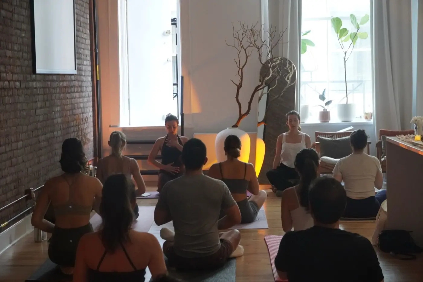 a seated circle of guests on mats facing two practitioners by tall windows, at the start of a pilates and rituals evening in a SoHo living room, New York