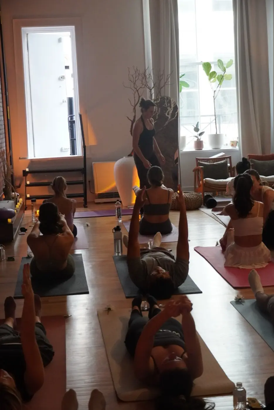 guests lying on mats with arms raised overhead as a practitioner walks between them, during a pilates class in a SoHo living room, New York