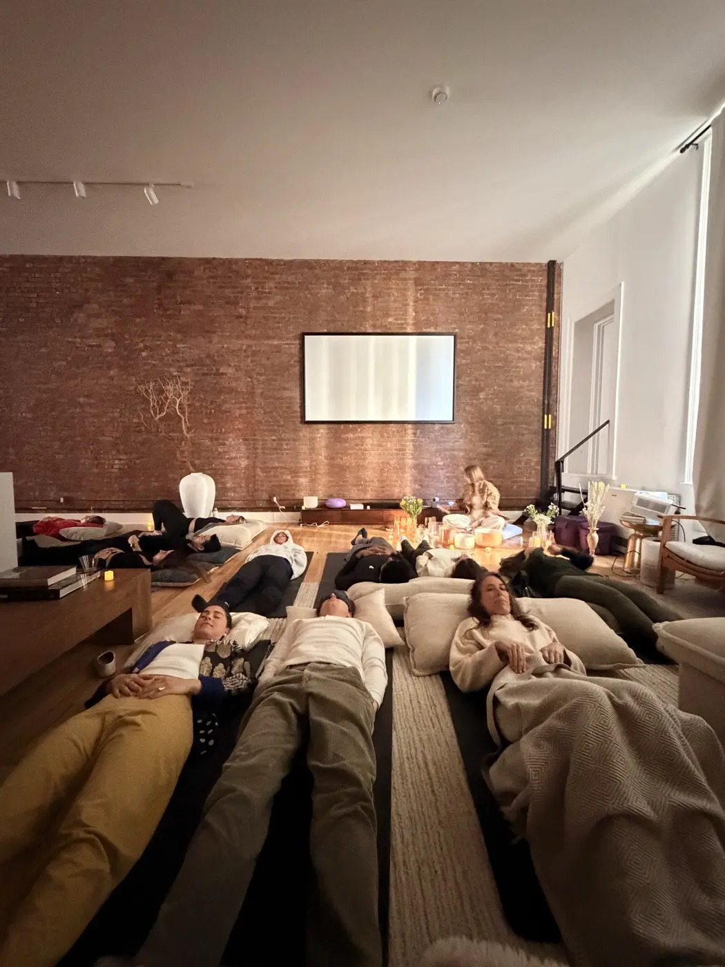 guests lying under blankets on the floor while a practitioner plays crystal bowls, during a sound bath in a SoHo living room, New York