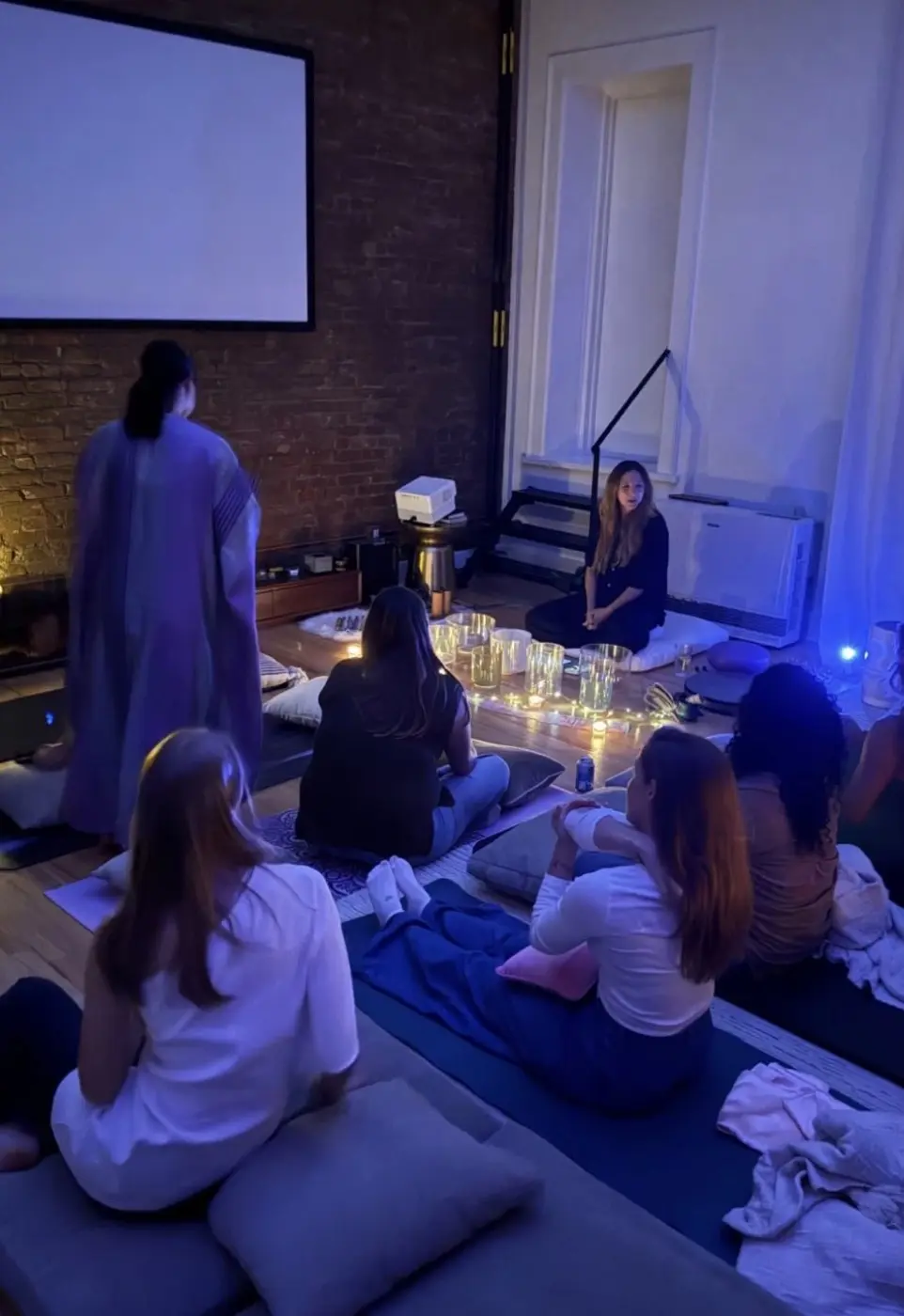 guests sitting on cushions in blue light around a ring of candles and crystal bowls, during an evening sound bath in a SoHo living room, New York