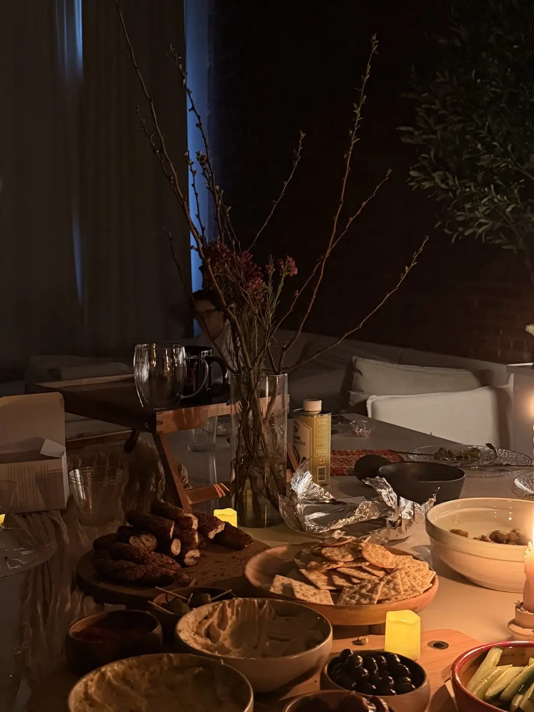 the food table at dusk in blue light, with hummus, crackers, olives and blossom branches in a glass vase, before a sound bath in a SoHo living room, New York